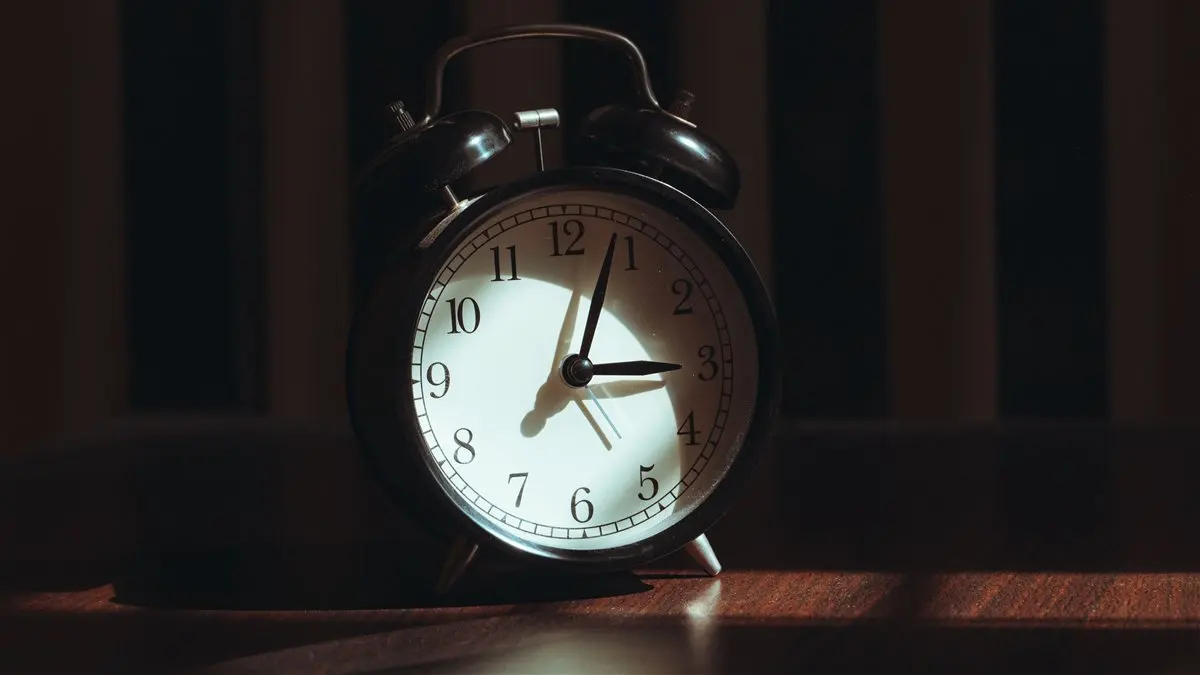 Bedside clock showing 3:12 a.m. in a dark bedroom with a lamp switched on