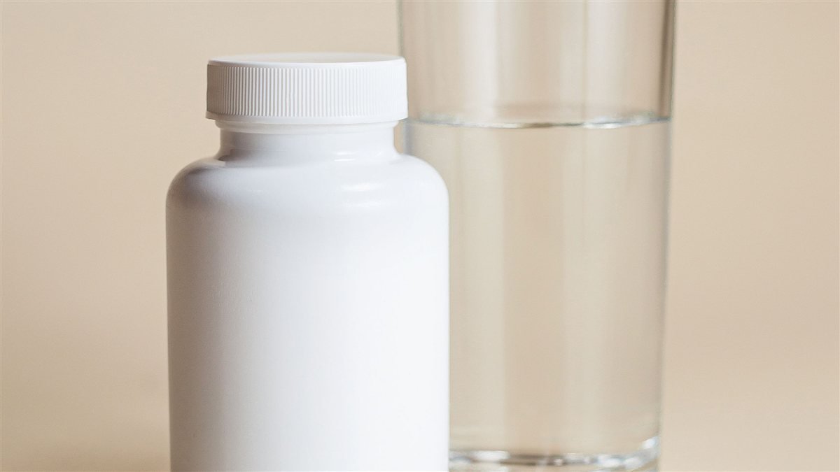 Unlabeled white supplement bottle beside a glass of water and capsules on a neutral background